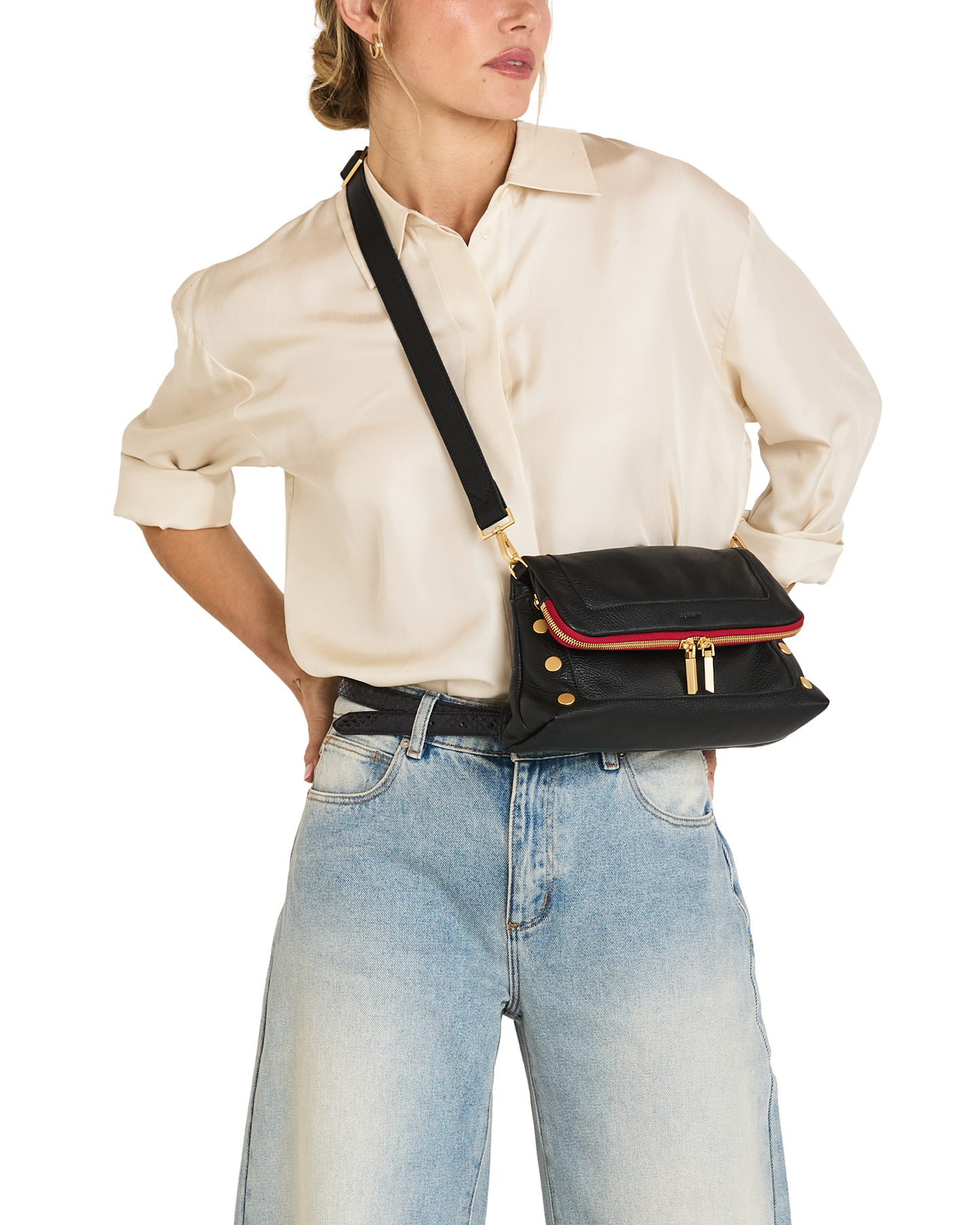 A model wearing a cream satin shirt and light blue jeans carries a black leather crossbody bag with a red leather trim and gold-tone hardware.