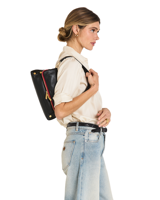 A woman in a cream shirt and light blue jeans shown from the side, wearing a black leather shoulder bag with gold hardware and a red accent stripe against a plain white background.