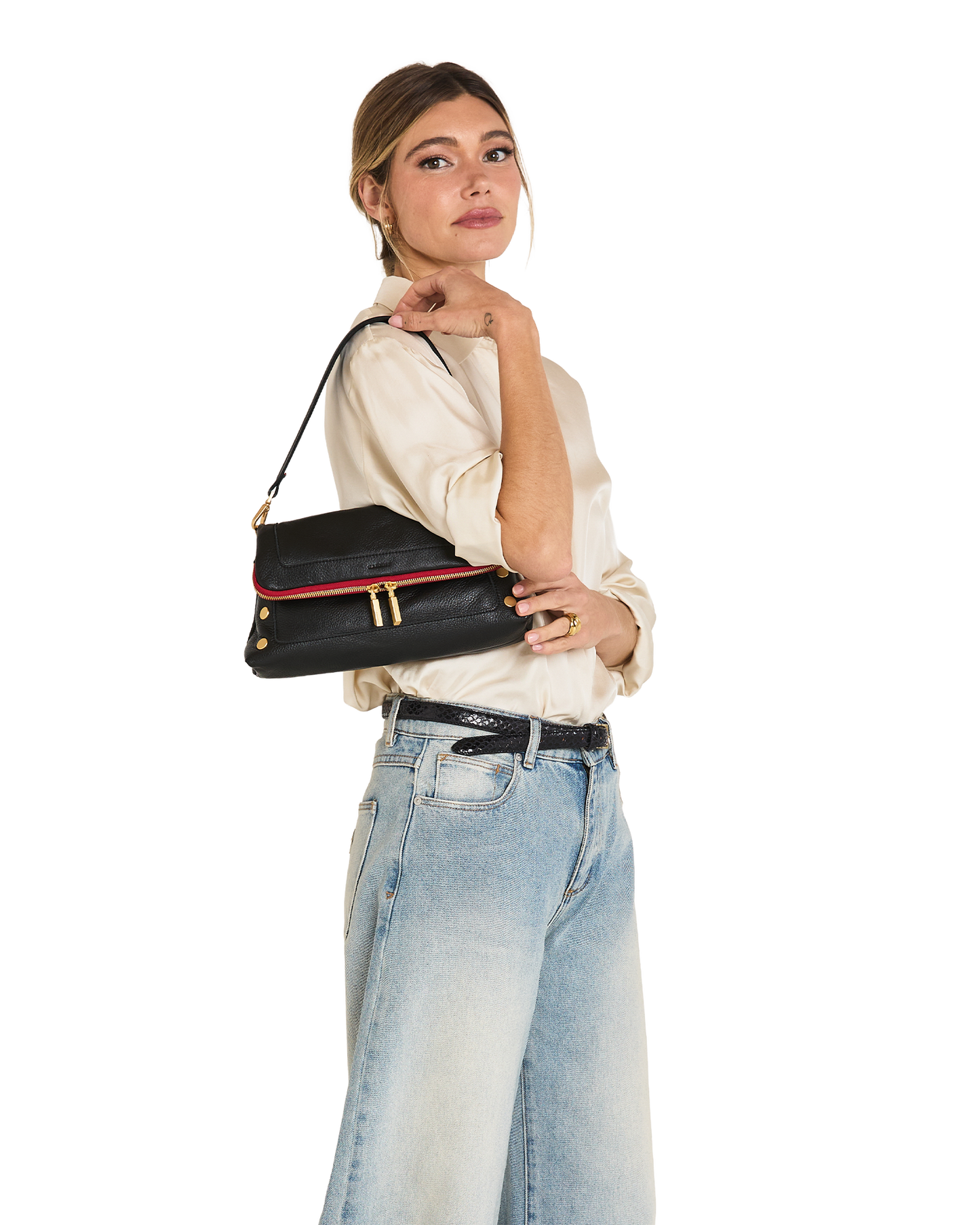 A woman wearing a cream blouse and light blue jeans carries a black shoulder bag with a red trim and gold hardware against a plain white background.