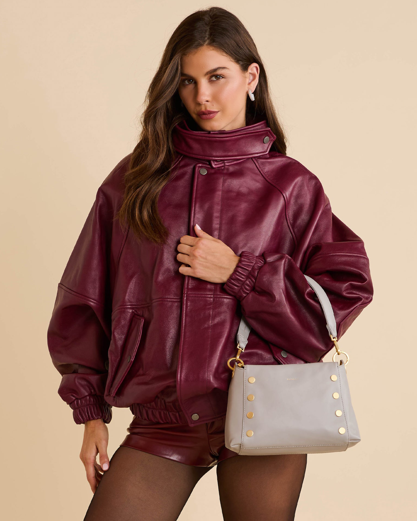 A woman models an oversized, shiny burgundy faux-leather jacket with a high collar, paired with matching burgundy shorts and a light gray shoulder bag with gold studs, against a solid beige background.