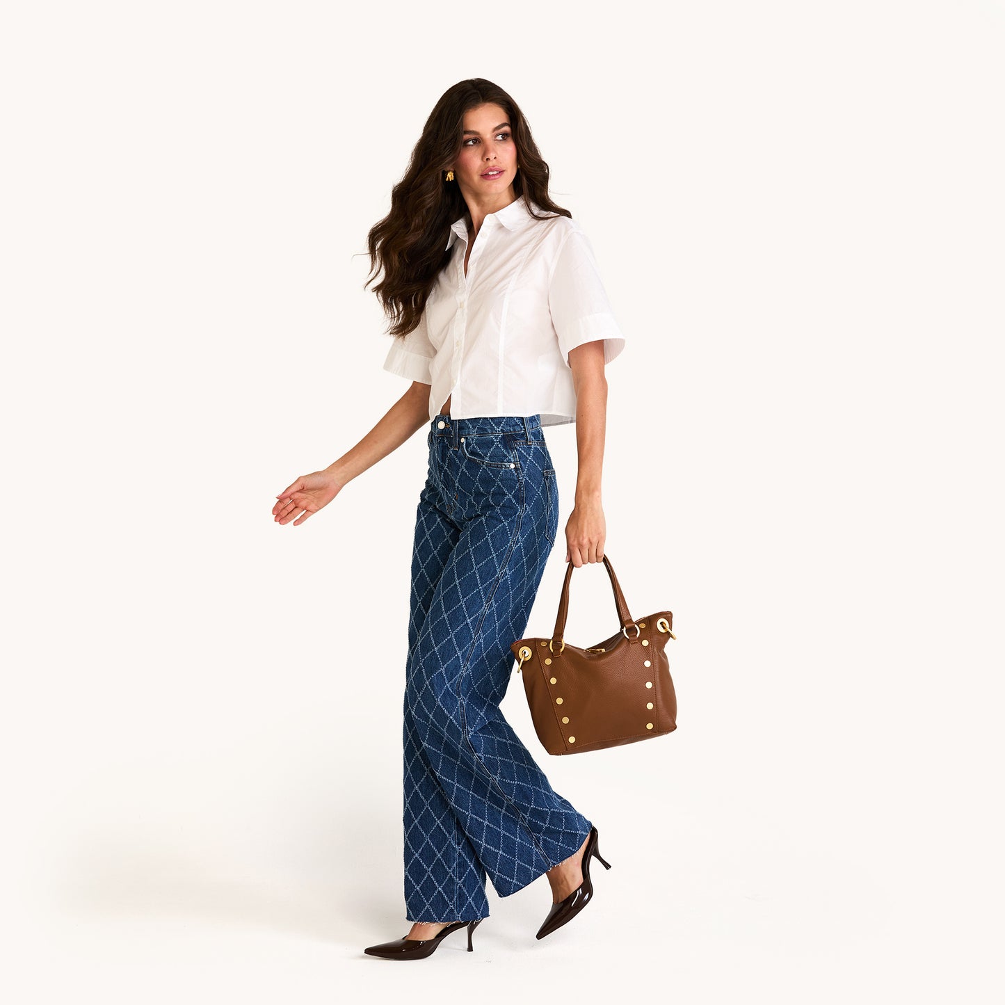 Woman wearing a white blouse and blue patterned pants, holding a brown handbag on a white background