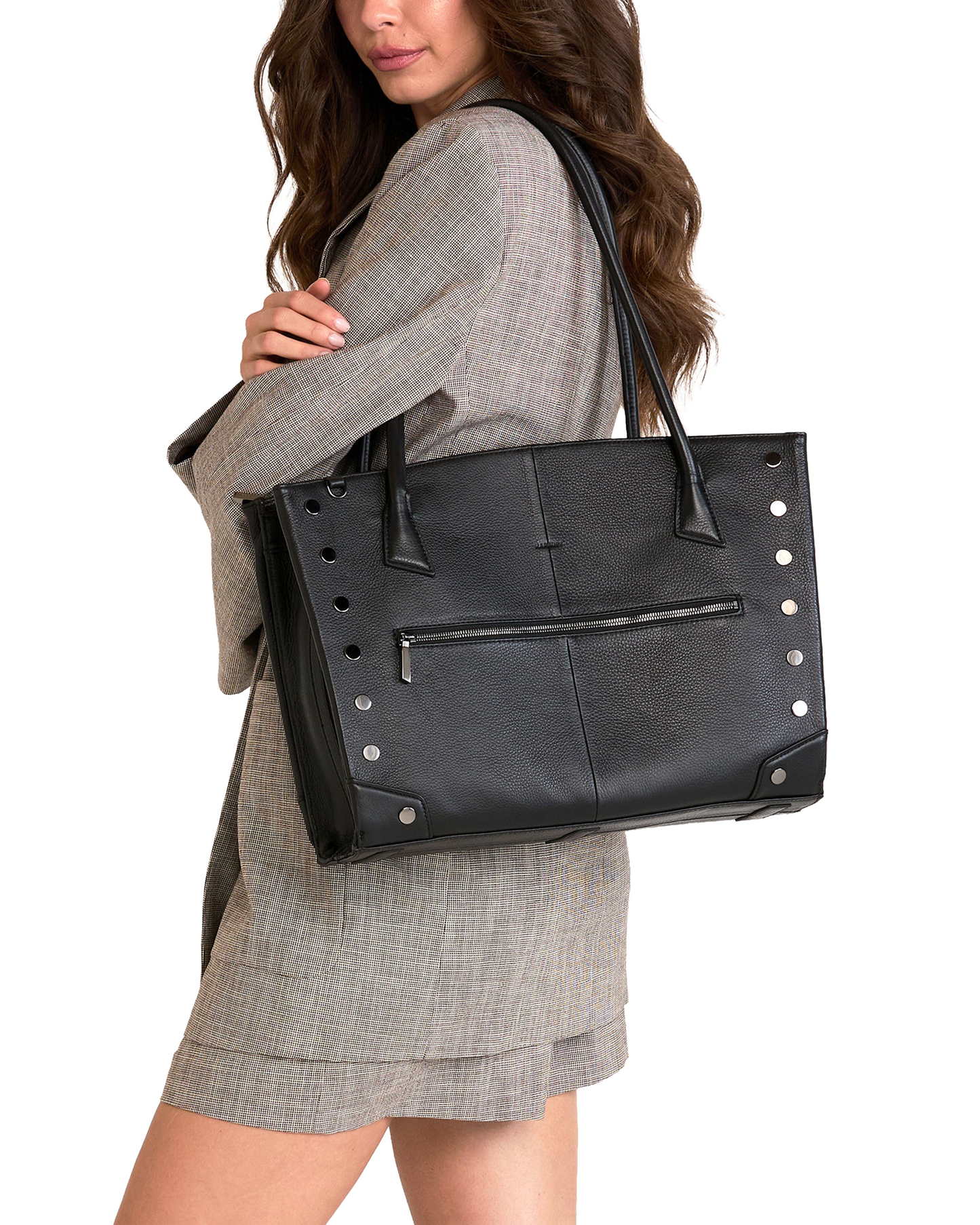 A woman wearing a gray plaid blazer and matching skirt carries a large black leather tote bag with silver-tone studs and a front zip pocket against a plain white background.