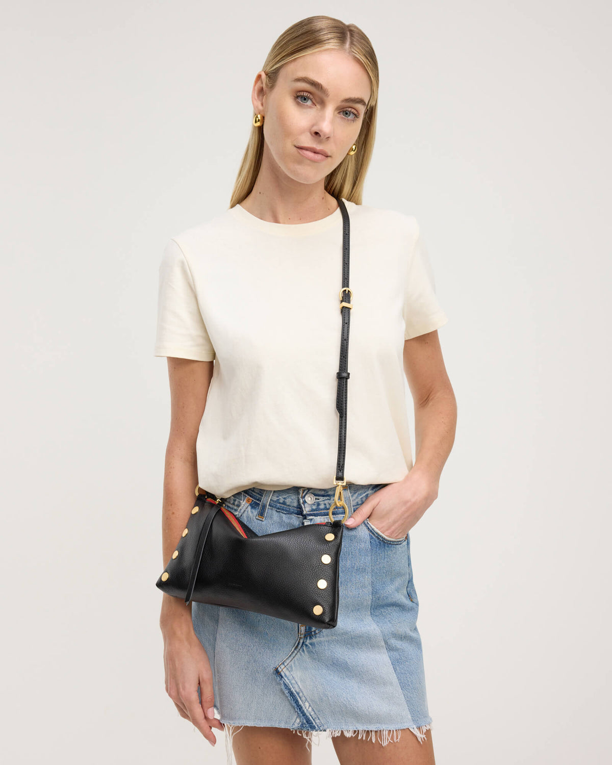 A woman modeling a black faux-leather crossbody bag with gold-tone studs and a gold buckle, styled with a cream t-shirt and denim skirt against a light gray studio background.