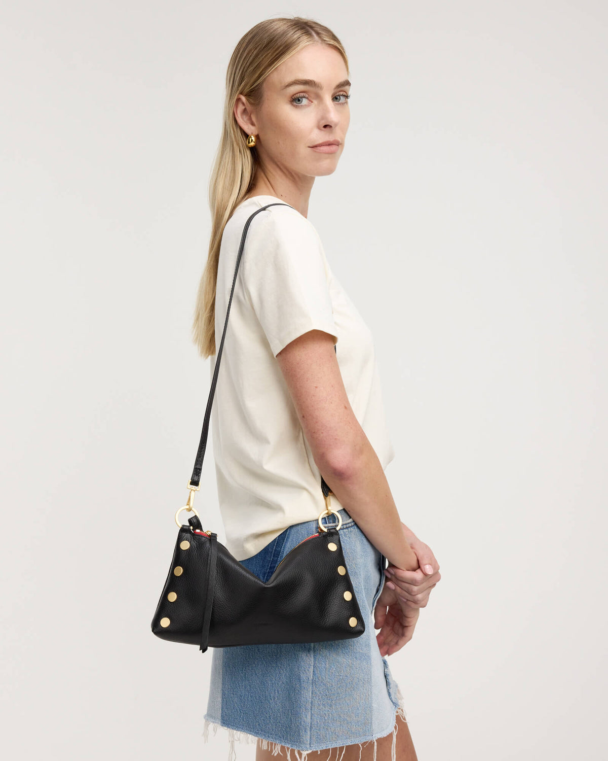 A woman in a white t-shirt and denim skirt models a black leather shoulder bag with gold studs and hardware against a plain light gray background.