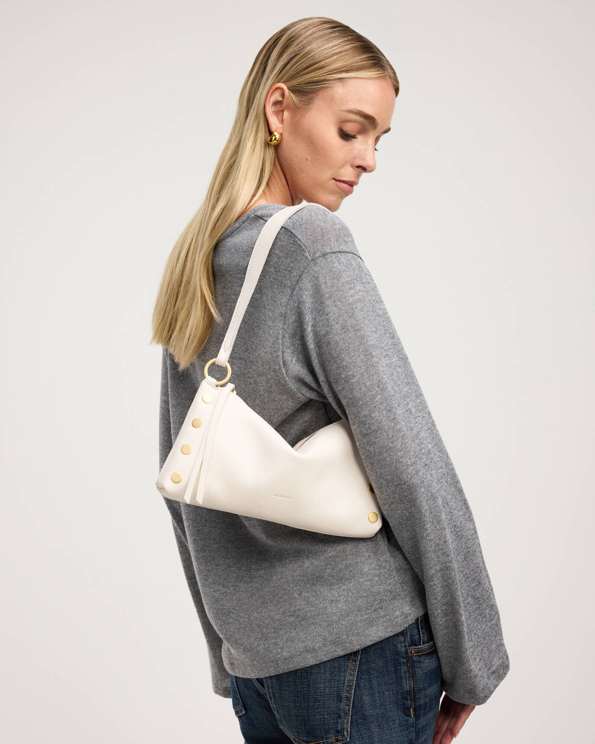 A blonde woman wearing a gray knit sweater and blue jeans, shown from a side-back angle while wearing a cream-colored slouchy shoulder bag with gold-tone hardware and decorative gold buttons.