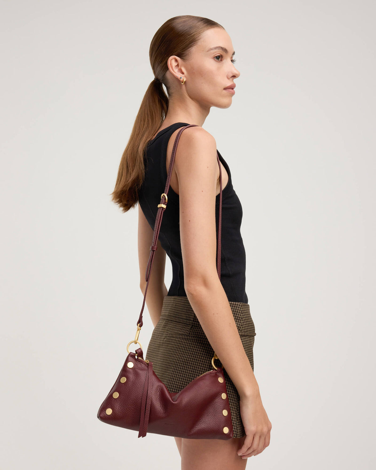 A woman in a black sleeveless top and brown patterned skirt is shown from the back and side profile, wearing a burgundy leather shoulder bag with gold-tone studs and a matching shoulder strap against a light gray studio background.
