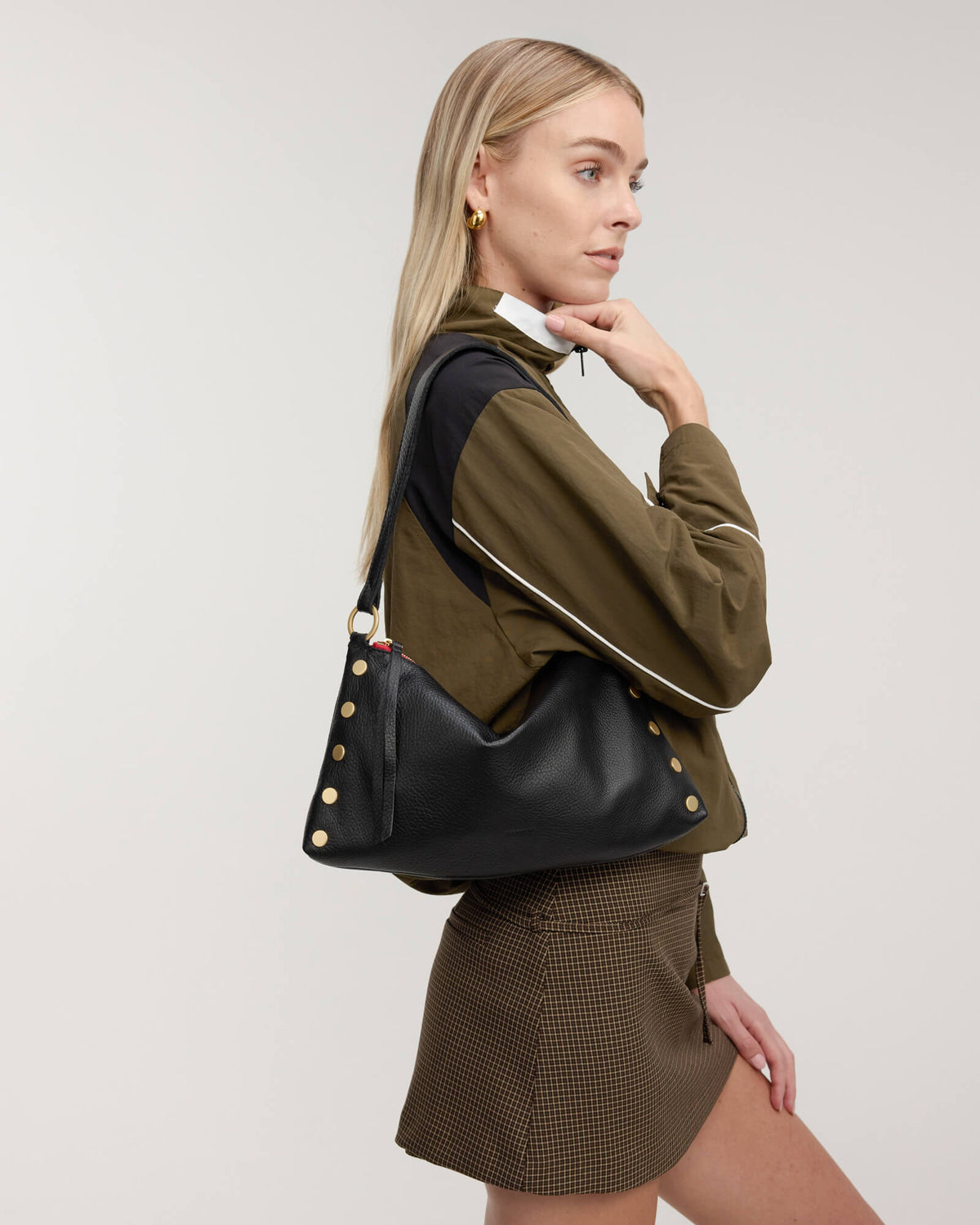 A blonde model wearing an olive green jacket and brown mini skirt, carrying a black leather shoulder bag with gold studs and a black strap against a light gray background.
