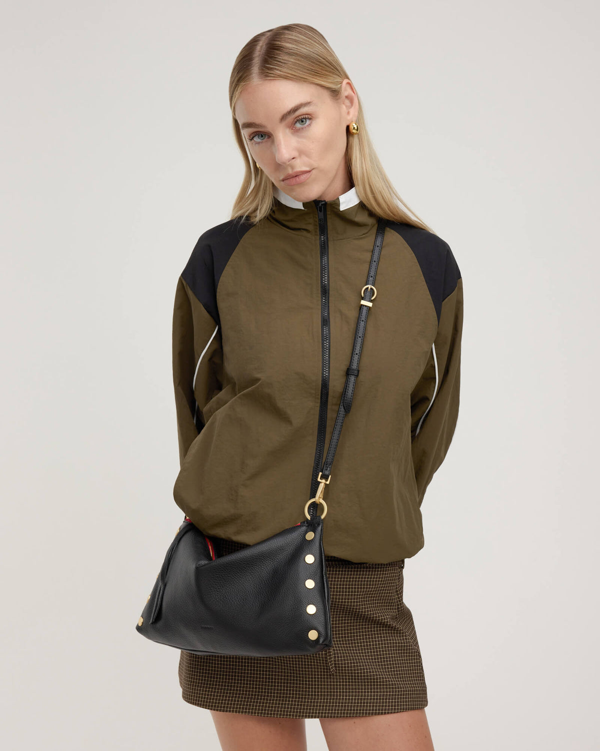 A model wearing an olive green and black color-blocked zip-up jacket, styled with a brown mini skirt and a black crossbody bag with gold hardware, against a light gray studio background.