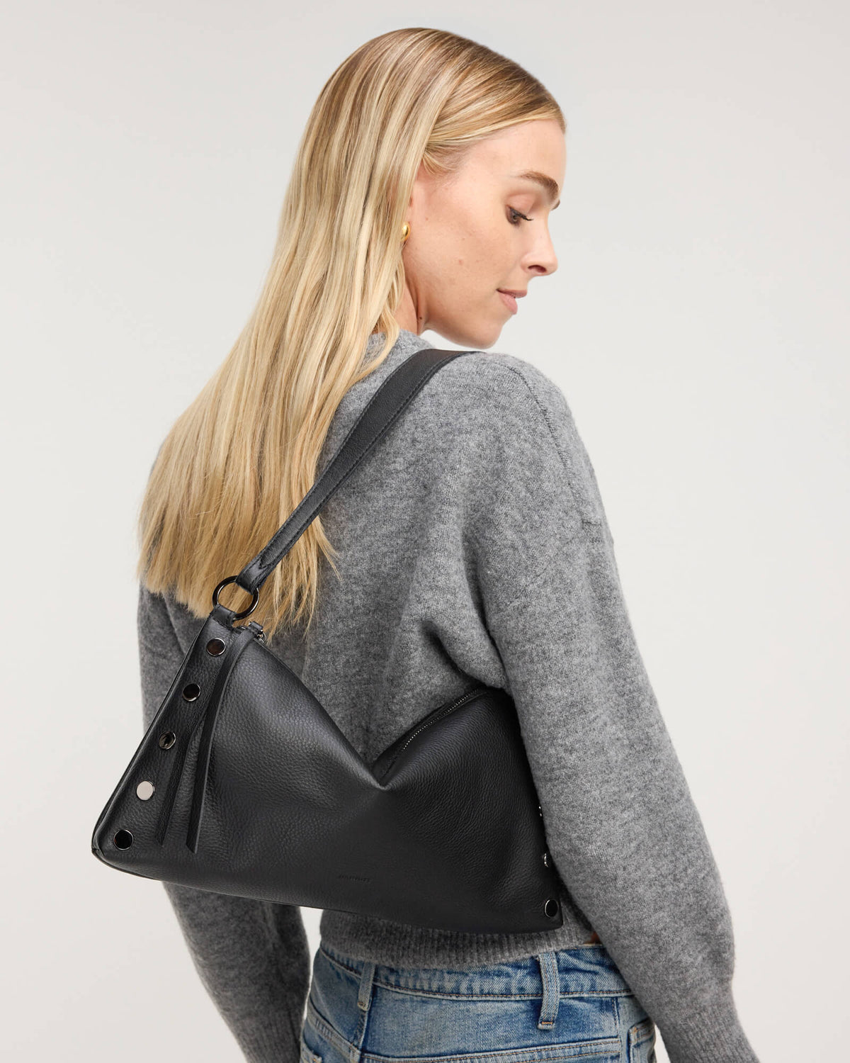 A blonde woman wearing a gray knit sweater and blue jeans, shown from a side-back angle while wearing a black slouchy shoulder bag with silver studs and a black strap against a light gray studio background.
