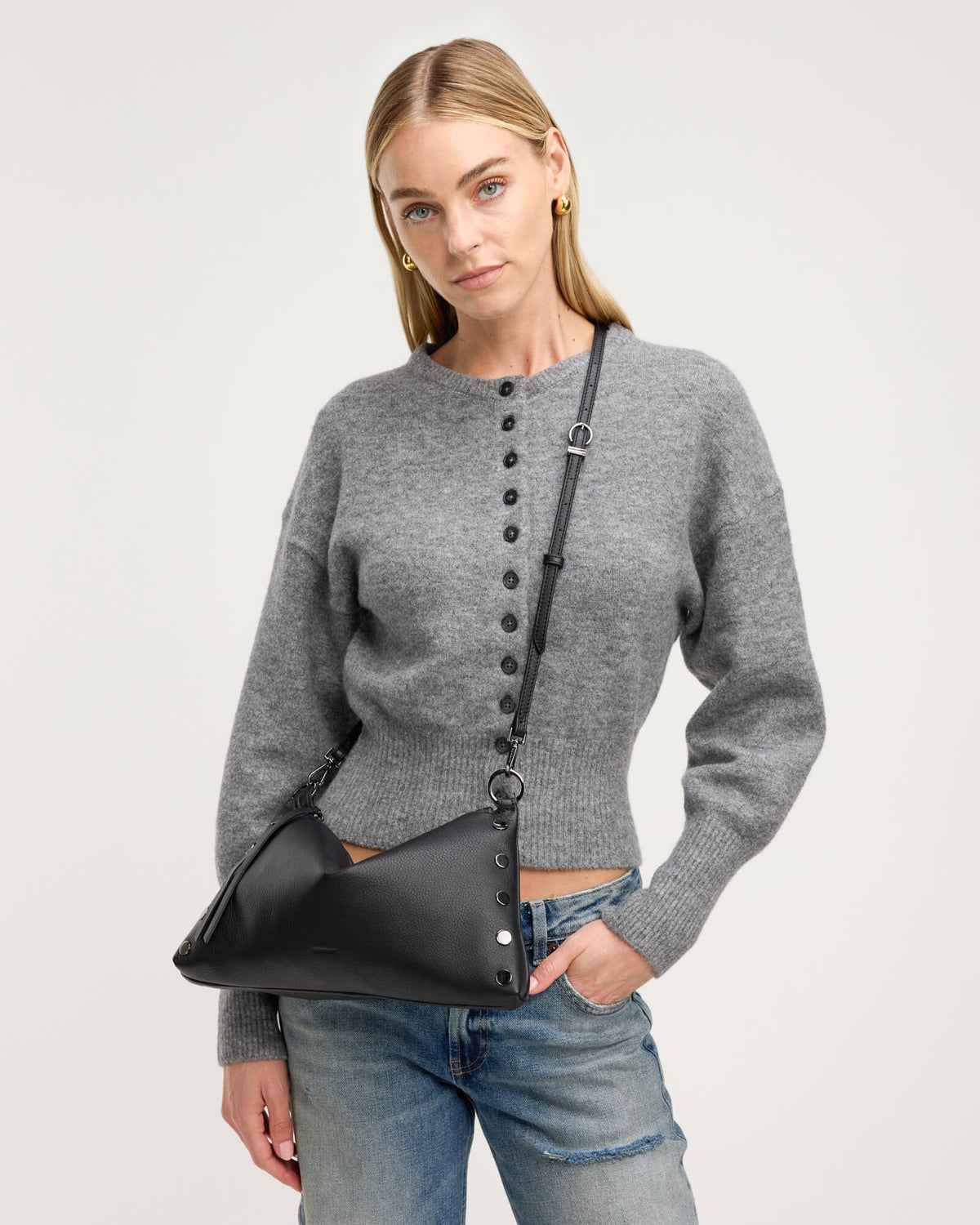 A blonde model wearing a gray button-front knit sweater and blue jeans, carrying a black crescent-shaped shoulder bag with a black strap and silver-tone hardware against a light gray studio background.