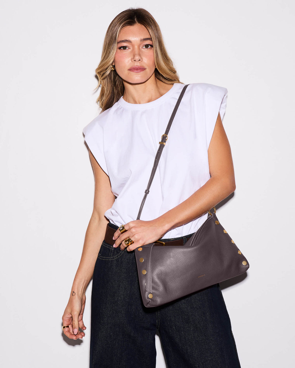 A woman wearing a white sleeveless top and dark jeans models a brown leather shoulder bag with gold-tone studs and hardware against a plain white studio background.