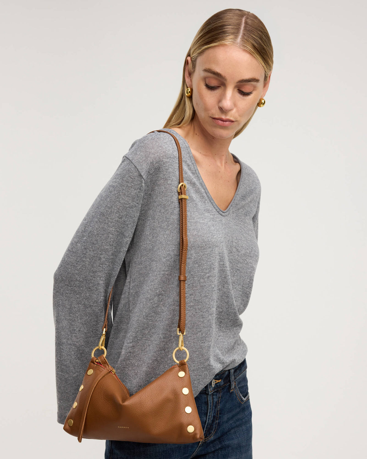 A woman wearing a gray long-sleeve V-neck top and blue jeans carries a brown leather shoulder bag with gold-tone hardware and decorative gold studs, photographed against a plain light gray studio background.