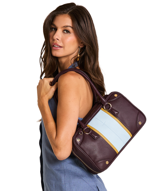 A model wearing a blue sleeveless top carries a structured leather handbag over her shoulder. The bag is deep burgundy with a wide light blue panel and a thin gold stripe detail.