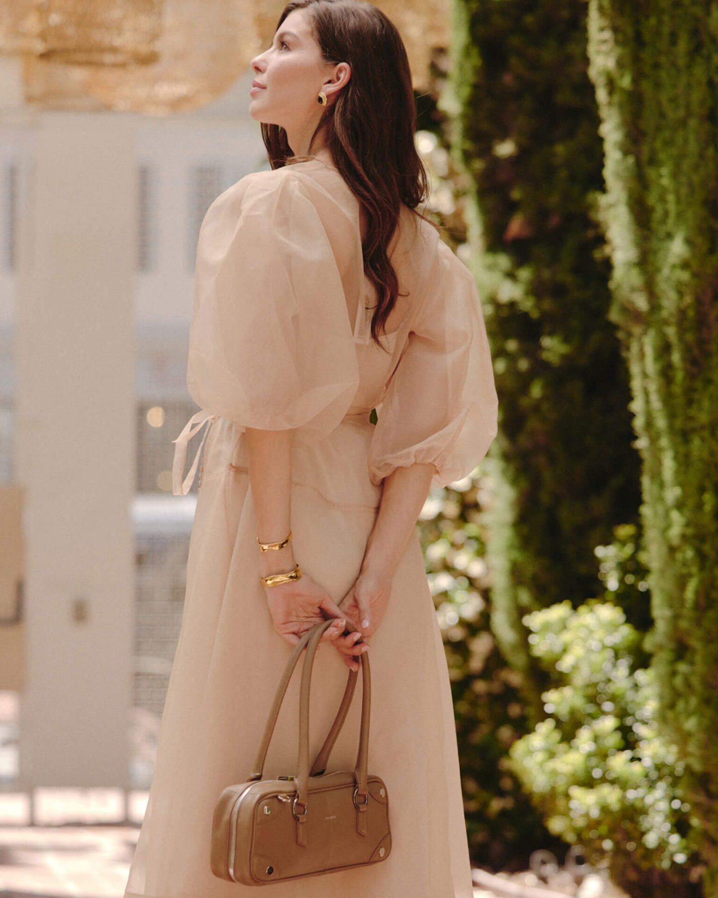 A woman wearing a pale blush pink puff-sleeve midi dress and holding a taupe leather handbag.