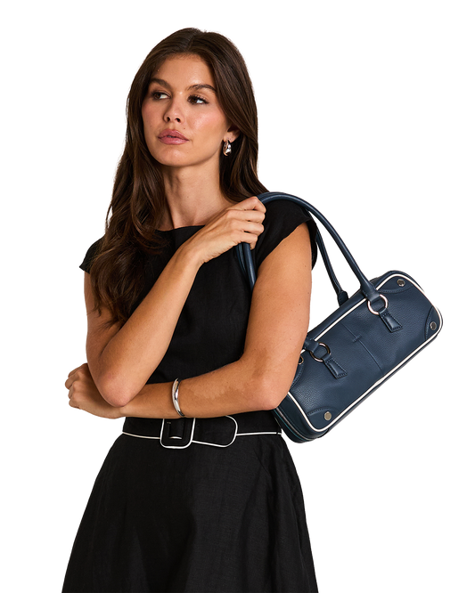 A woman in a black sleeveless top and skirt carries a navy blue leather handbag with white piping and silver hardware.