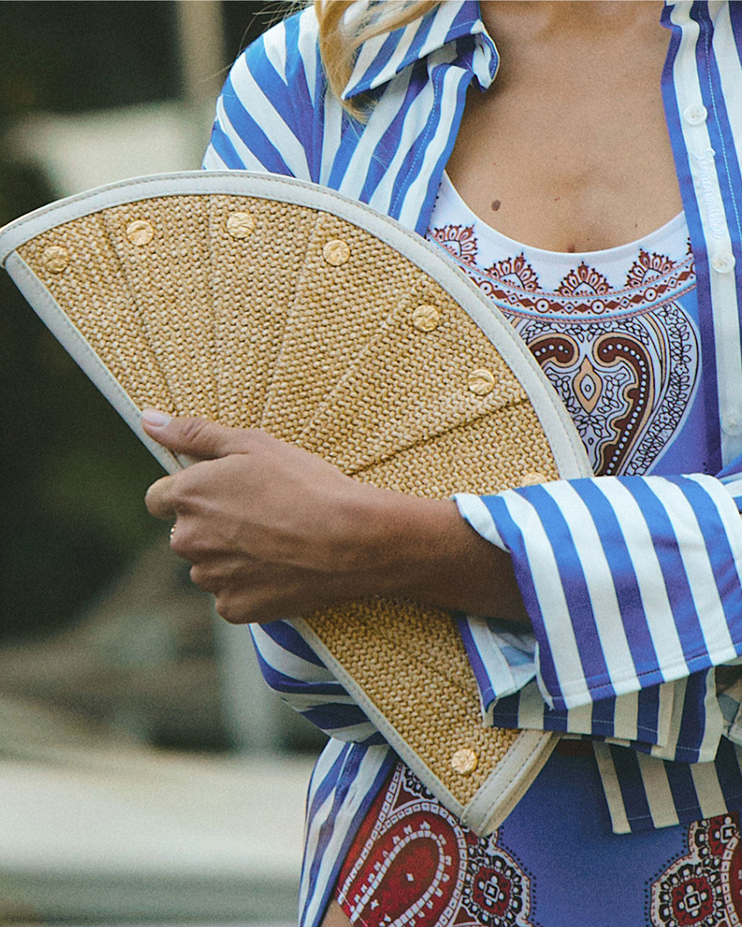 A close-up lifestyle shot of a person holding a woven straw folding fan. The person is wearing a blue and white striped button-up shirt over a white tank top with a red and blue paisley print.