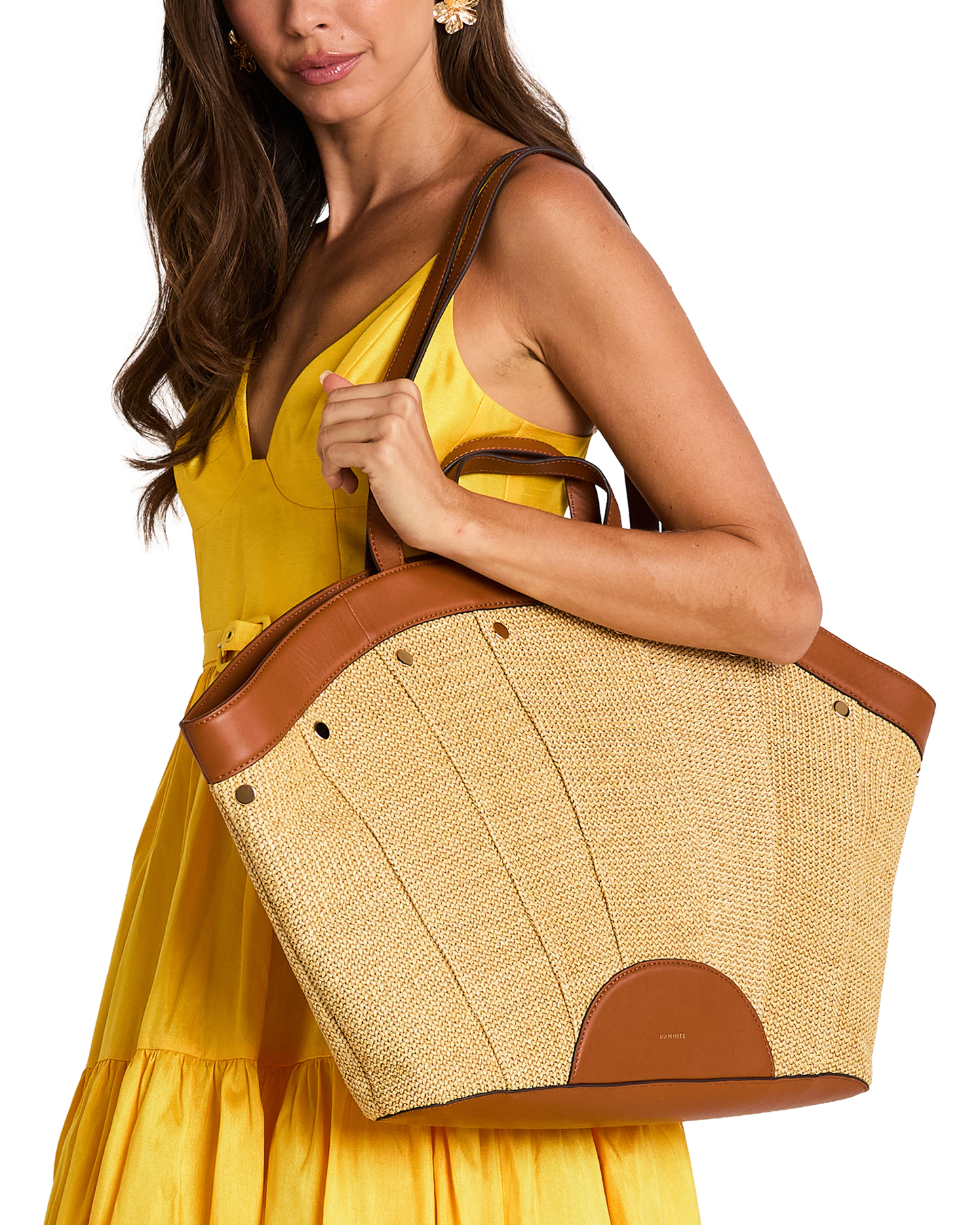 A woman in a yellow dress carries a large woven straw tote bag with brown leather trim and handles.
