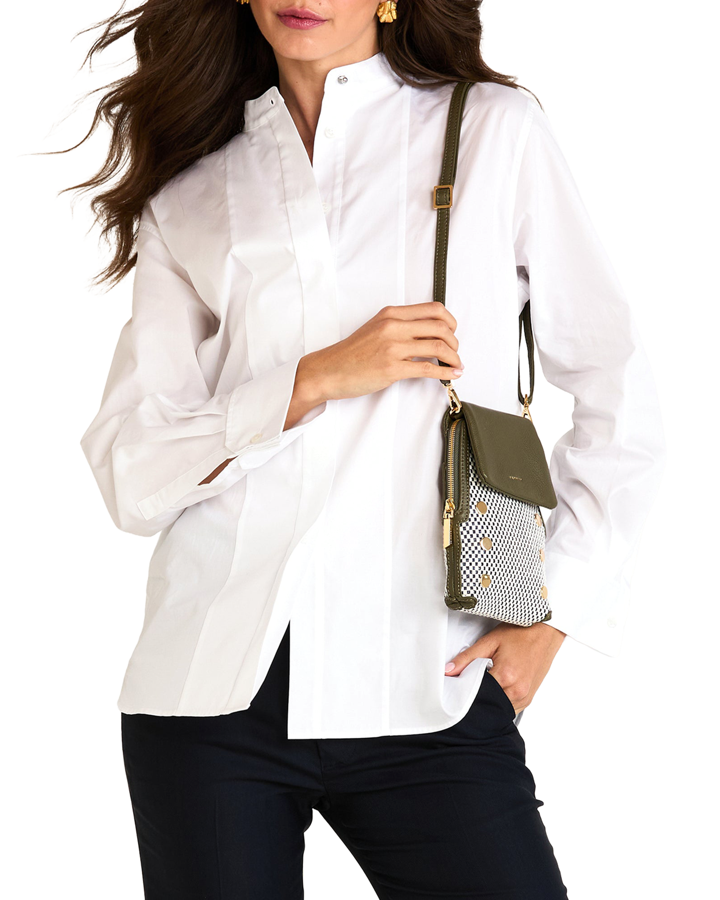 A woman wearing a white button-up blouse and dark pants, holding a small olive green and patterned crossbody bag against a plain white background.