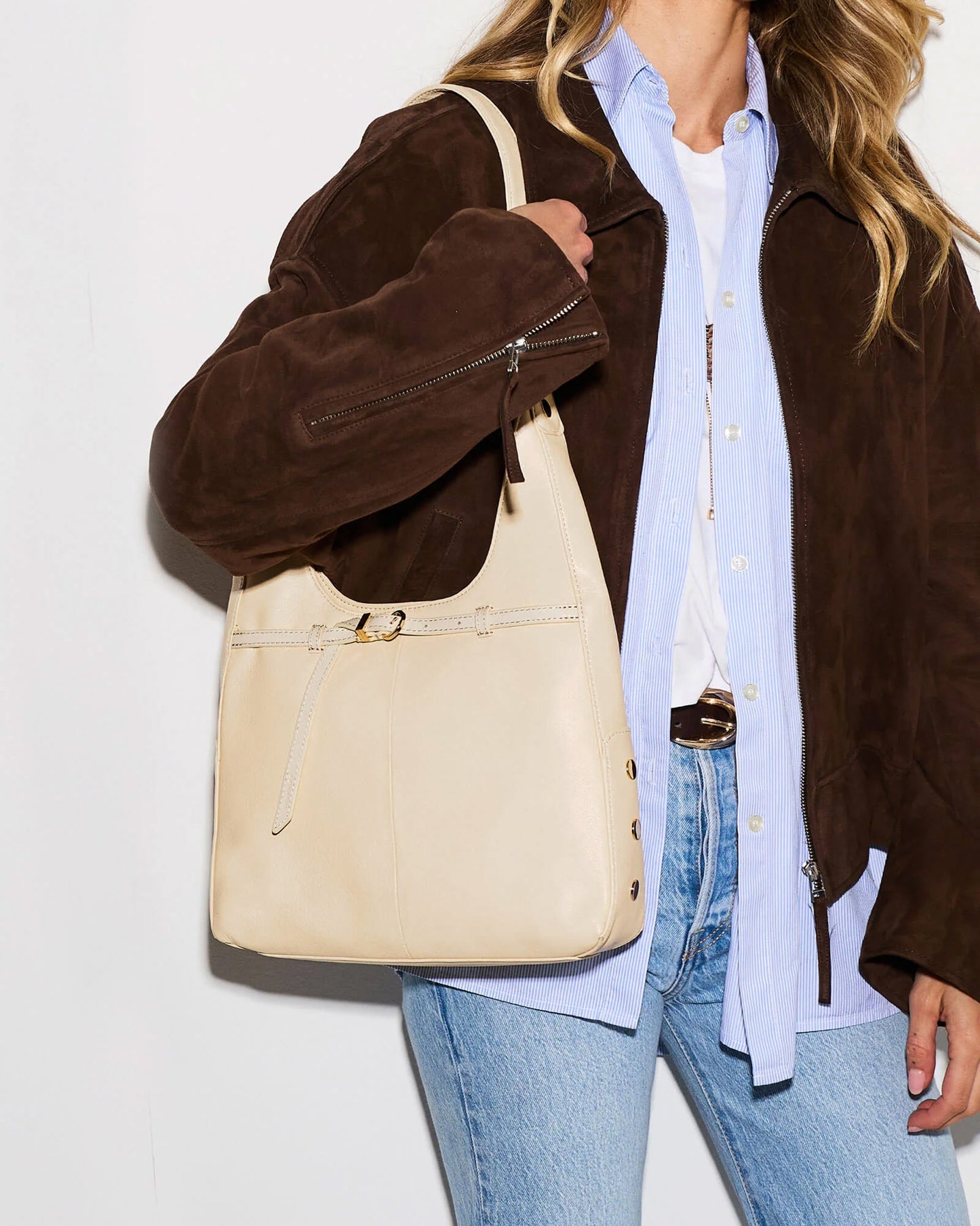 A woman wearing a brown suede jacket, light blue button-down shirt, white tee, and blue jeans carries a large cream-colored leather shoulder bag with gold-tone hardware.