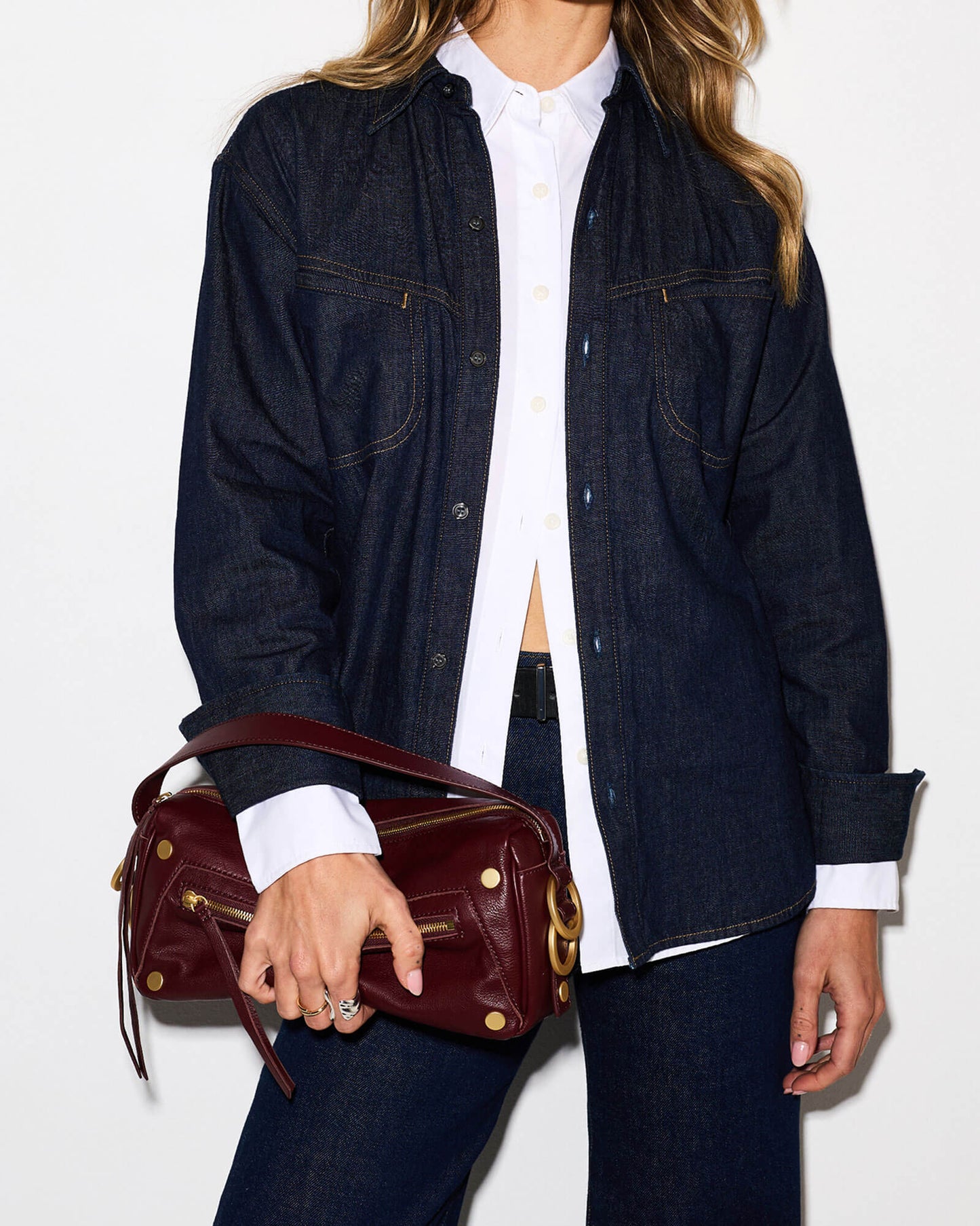 A model wearing a dark indigo denim button-up shirt over a white collared shirt, paired with dark blue jeans and holding a burgundy leather shoulder bag with gold hardware.