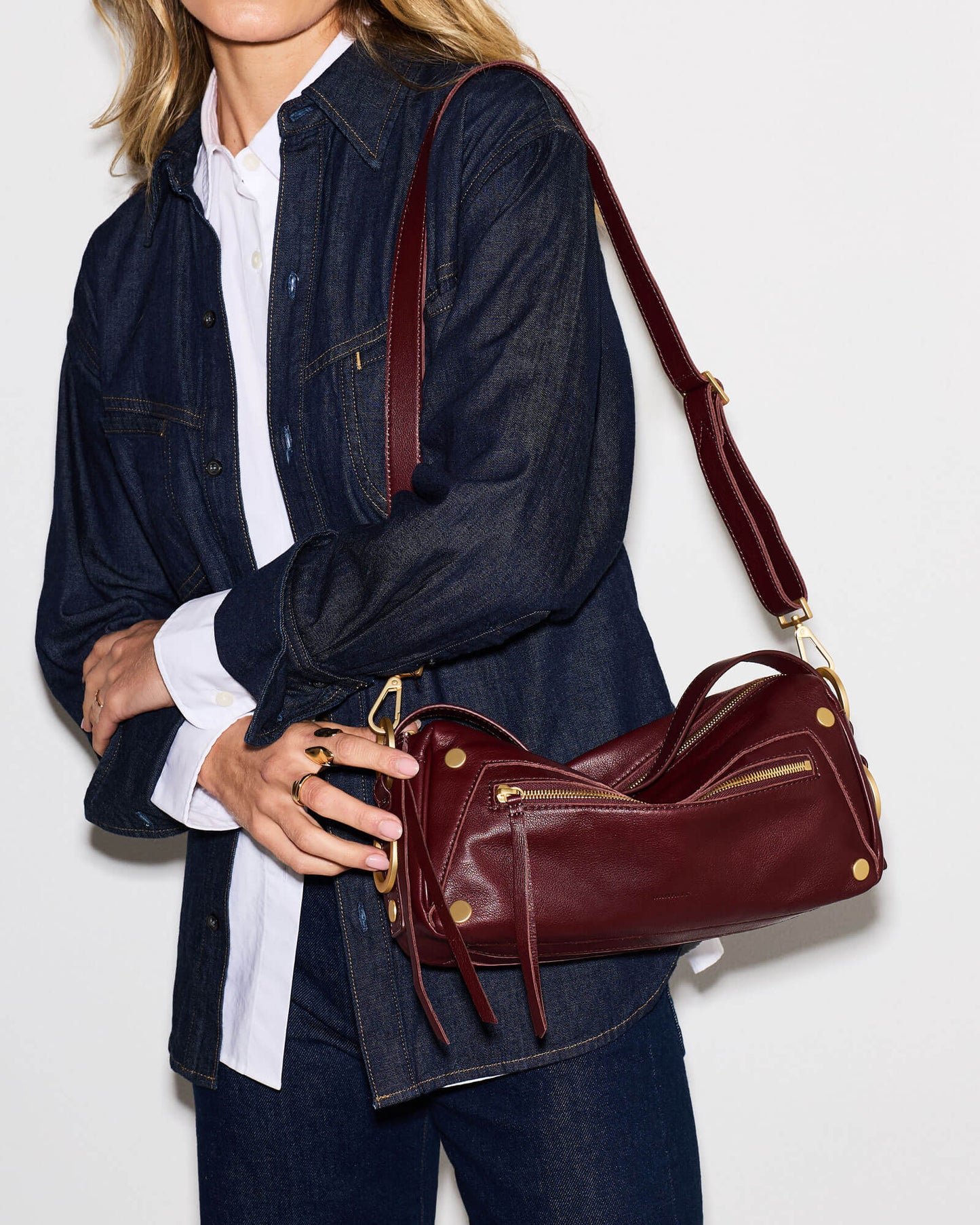 A woman wearing a dark denim shirt and white collared shirt carries a burgundy leather shoulder bag with gold-tone hardware against a plain white background.