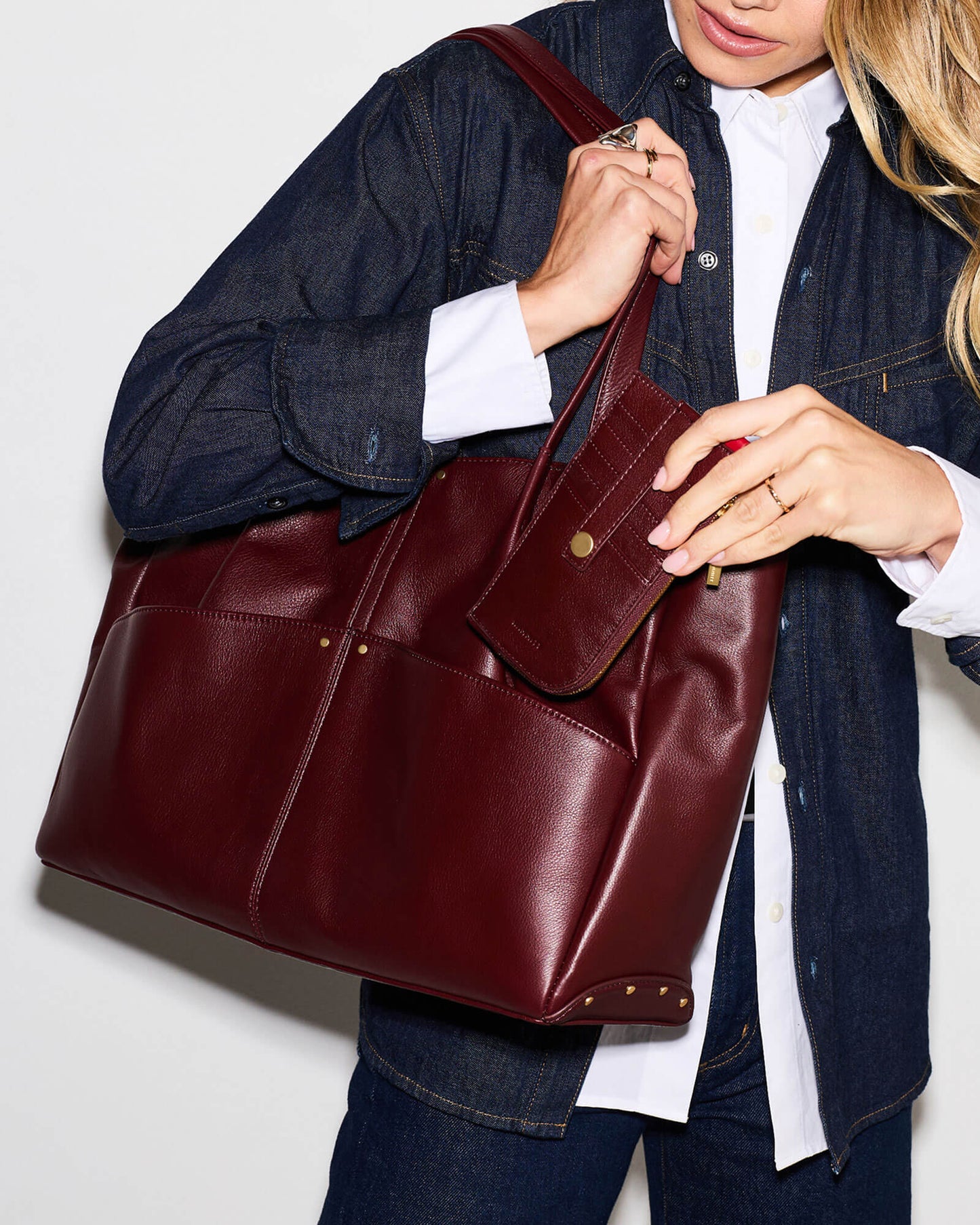 A woman wearing a dark denim jacket and white shirt carries a large burgundy leather tote bag with a matching zippered pouch, shown against a plain light background.