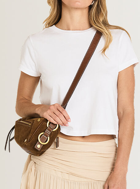 A woman wearing a white cropped t-shirt and beige pleated skirt carries a brown suede crossbody bag with a wide brown strap and gold-tone hardware.