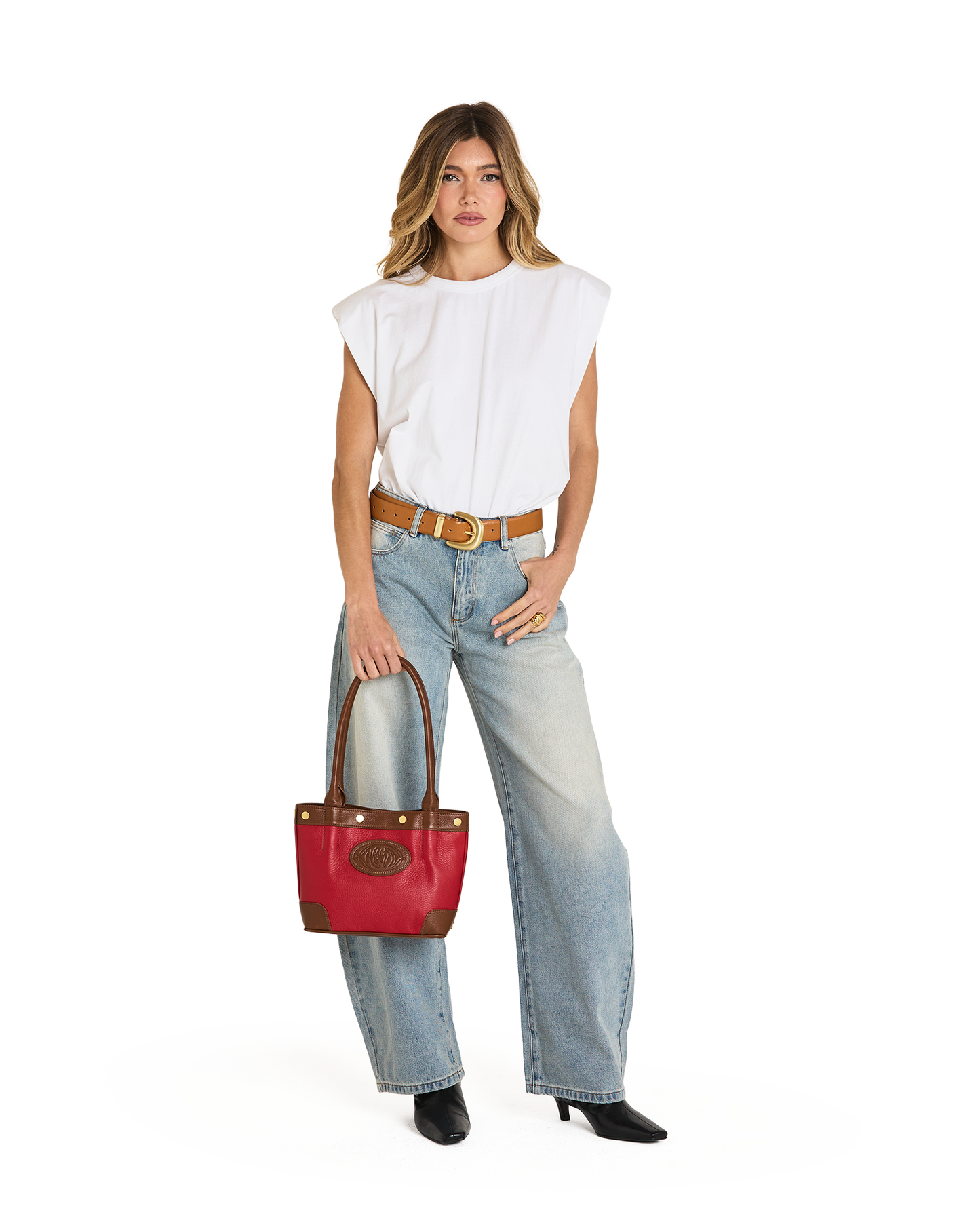 A woman modeling a white sleeveless top, light-wash wide-leg jeans, a brown belt, and black boots, holding a red leather handbag with brown leather handles and trim against a plain white background.