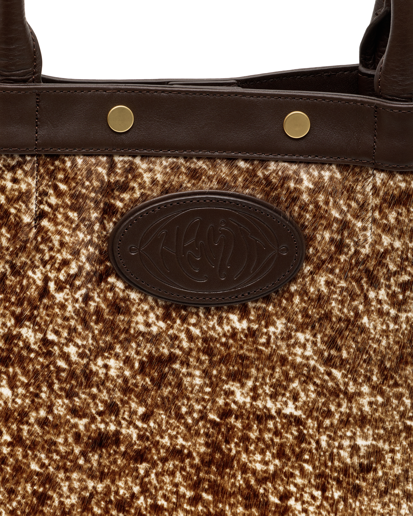 Close-up of a handbag featuring a brown and beige cowhide patterned body, dark brown leather trim with gold-tone rivets, and a dark brown oval embossed logo patch.