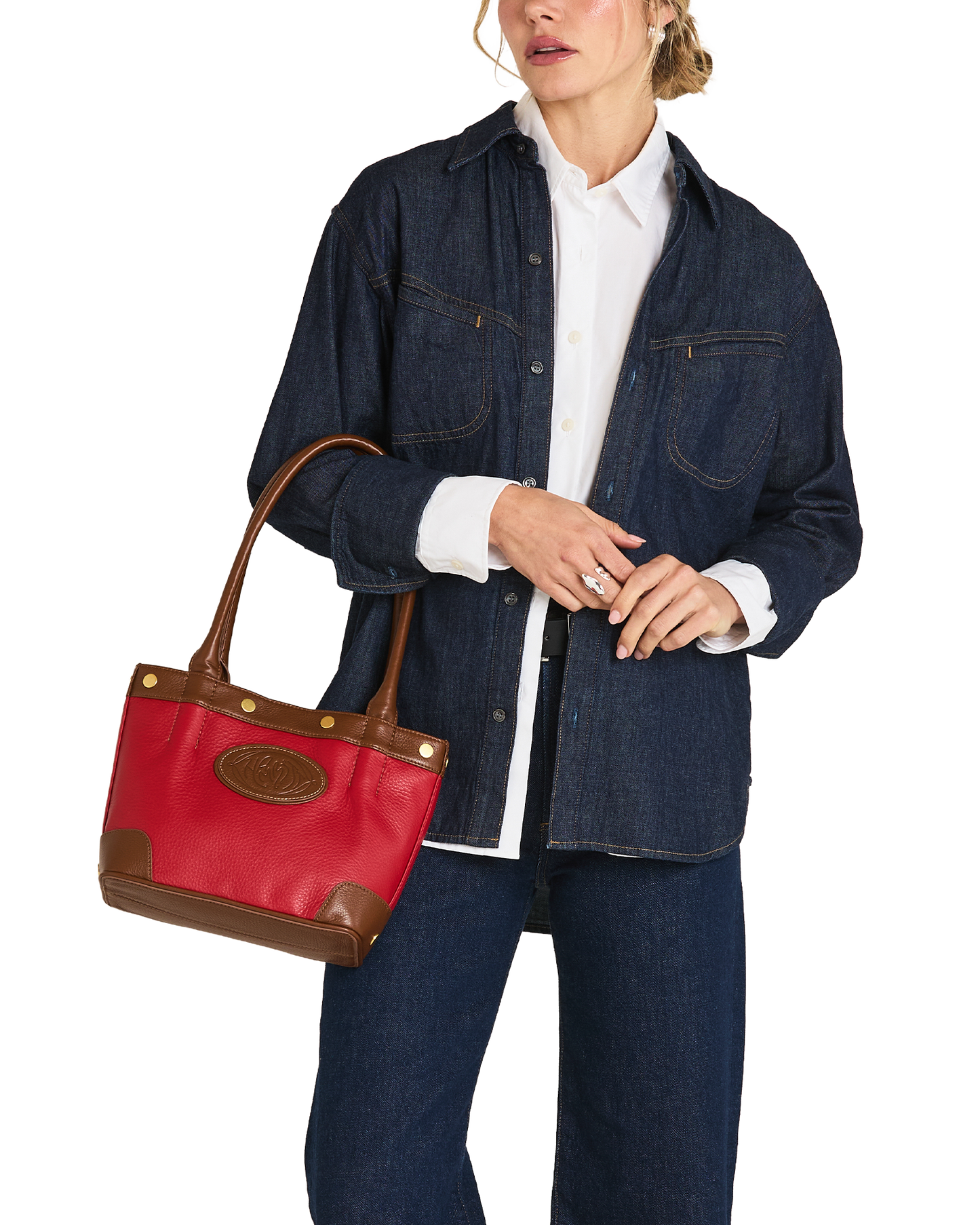 A model wearing a dark blue denim shirt jacket, white button-down shirt, and dark blue jeans, holding a red leather tote bag with brown leather handles and trim.