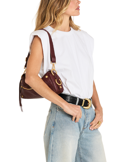 A woman wearing a white sleeveless top, light blue jeans, and a black belt with a gold buckle, carrying a dark burgundy leather shoulder bag with gold hardware.