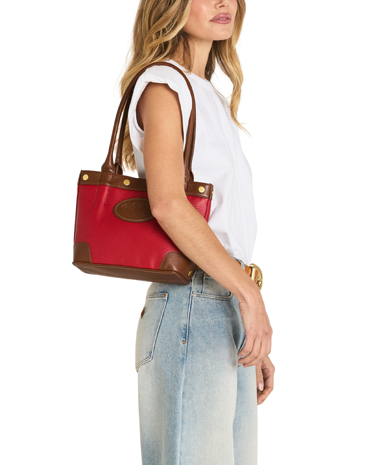 A woman in a white sleeveless top and light blue jeans carries a red and brown leather tote bag over her shoulder, shown against a plain white background.