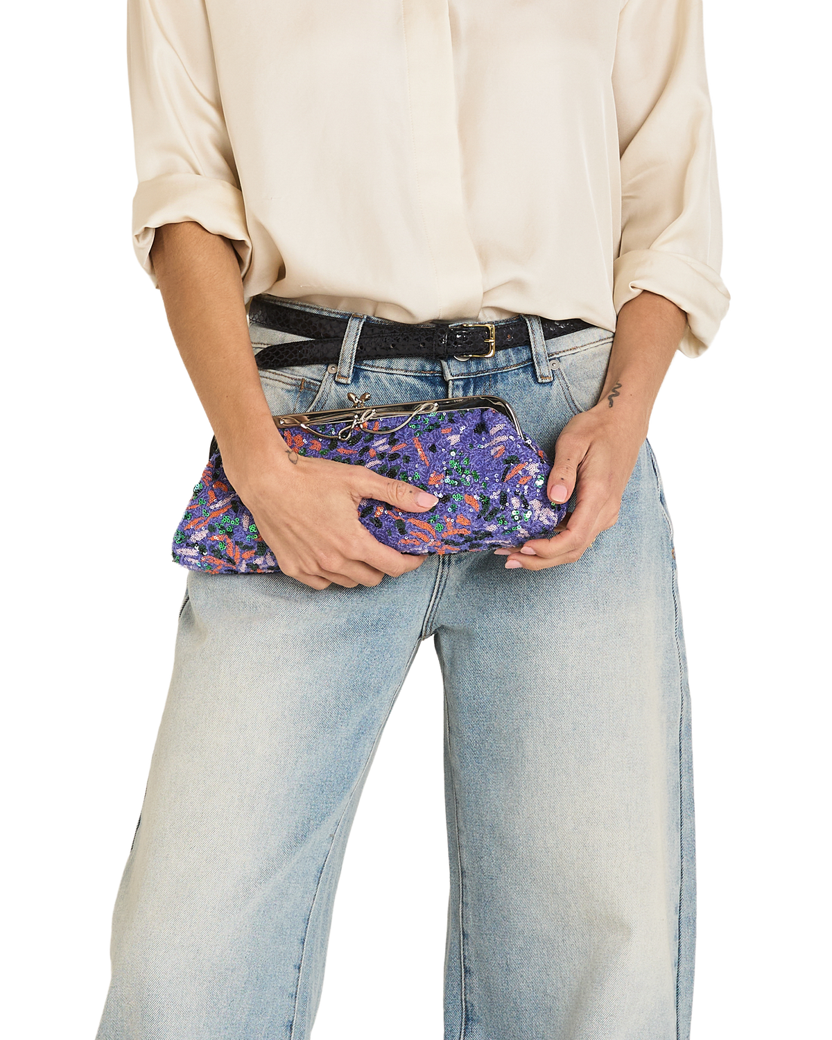 A person wearing a cream blouse, black belt, and light-wash wide-leg jeans holds a purple floral clutch bag against their hip against a plain white background.