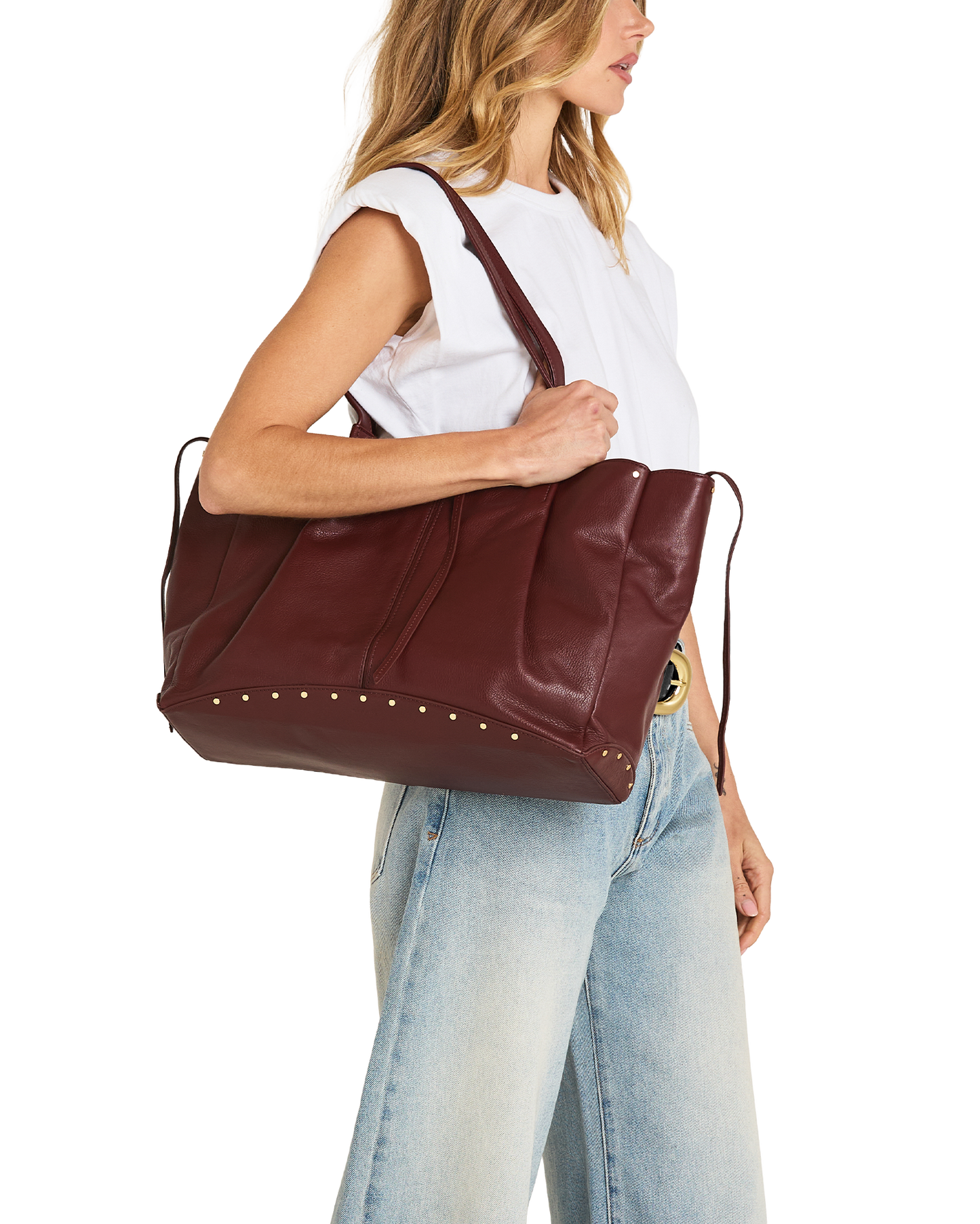 A woman wearing a white sleeveless top and light blue jeans carries a large burgundy leather tote bag with gold-tone studs and a shoulder strap against a plain white background.