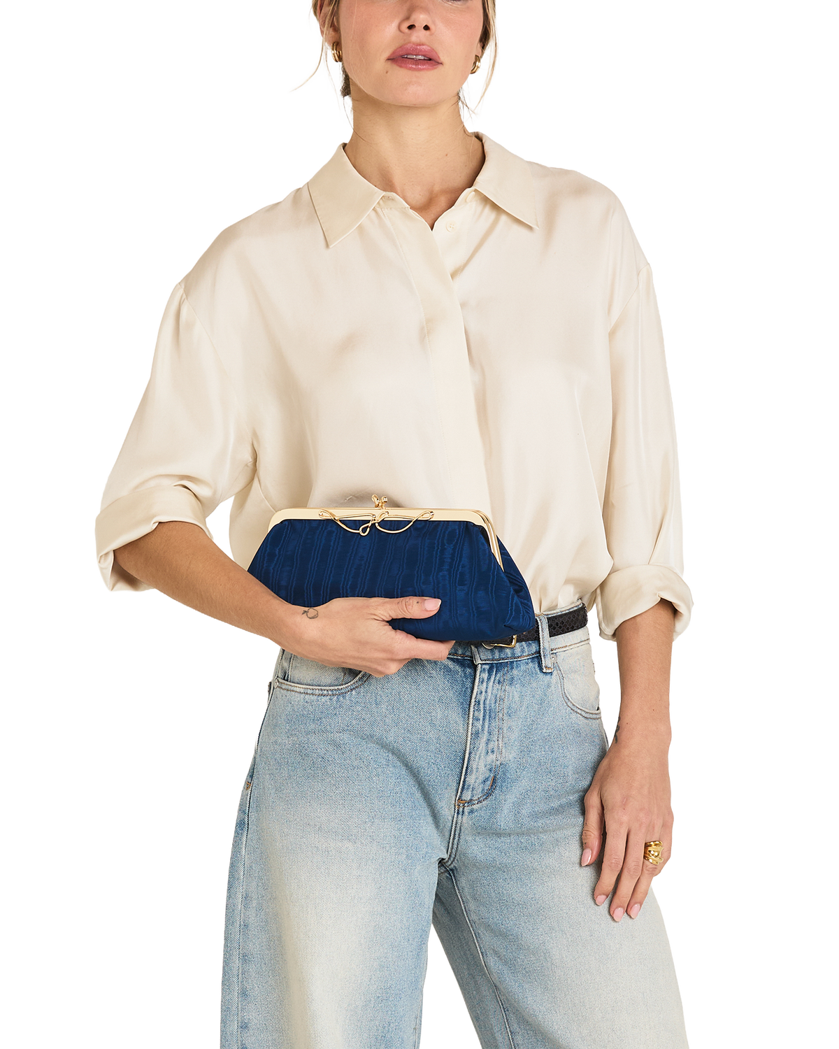 A model wearing a cream-colored silk shirt and light-wash blue jeans holds a navy blue velvet clutch with a gold-tone kiss-lock closure.