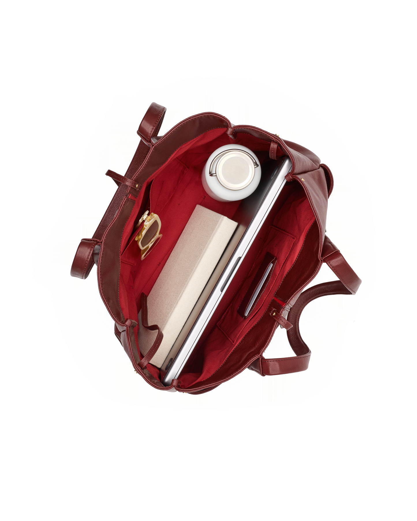 An overhead view of a dark red leather tote bag shown open, filled with a laptop, notebook, pen, and a white travel mug, on a solid white background.