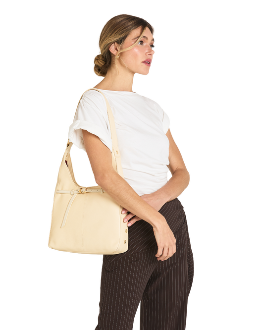 A woman wearing a white t-shirt and dark brown pinstriped trousers carries a light beige leather shoulder bag on her shoulder, shown from a side profile against a solid white background.