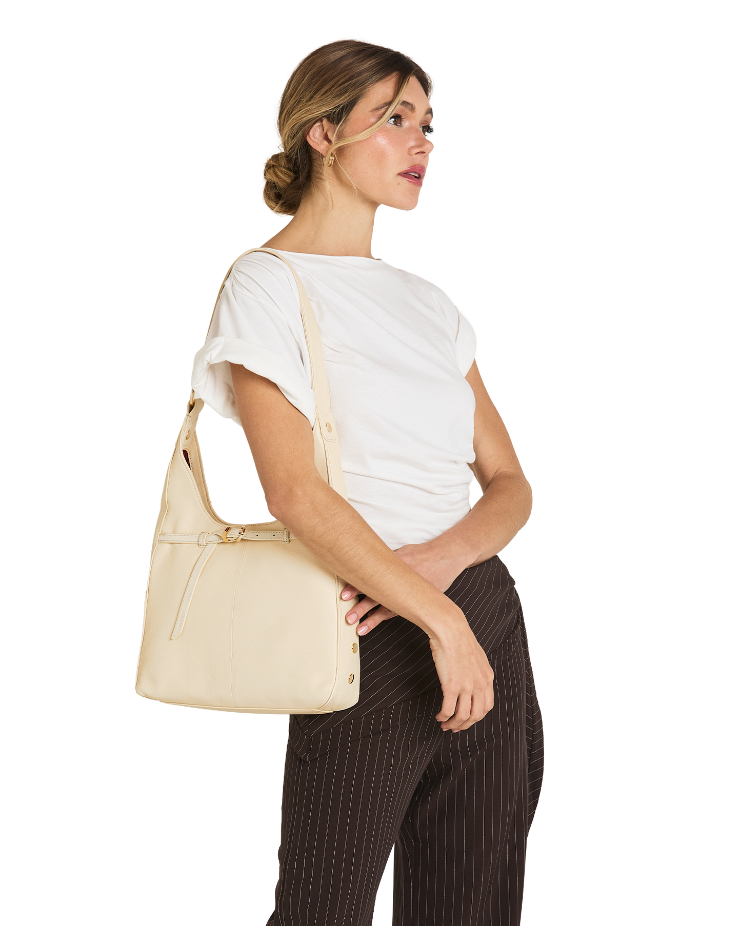 A woman wearing a white t-shirt and dark brown pinstriped trousers carries a light beige leather shoulder bag on her shoulder, shown from a side profile against a solid white background.