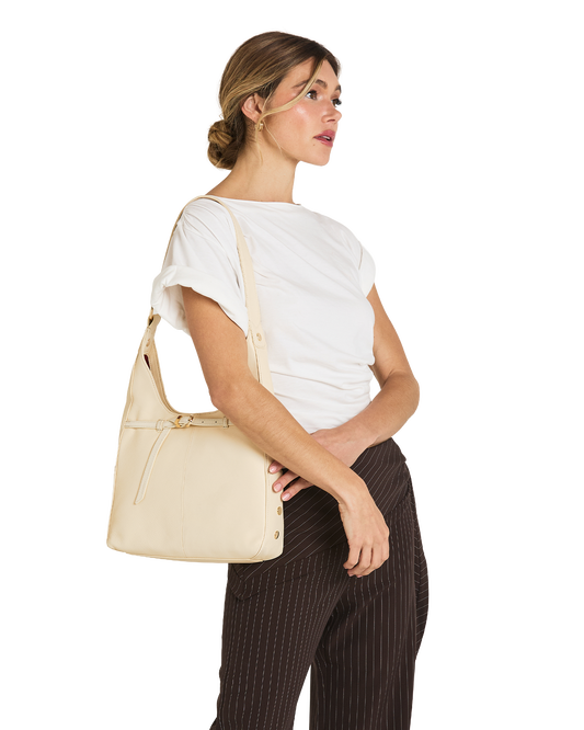A woman wearing a white t-shirt and dark brown pinstriped trousers carries a light beige leather shoulder bag on her shoulder, shown from a side profile against a solid white background.