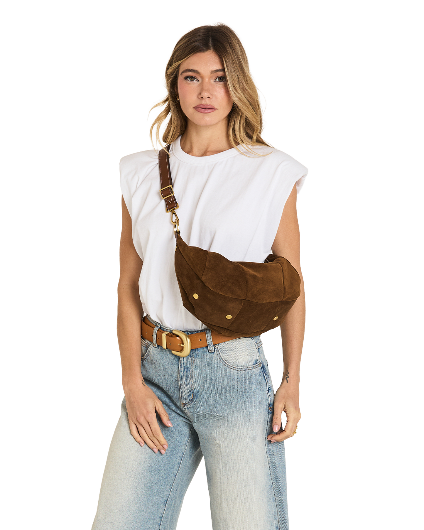 A woman wearing a brown suede crescent-shaped crossbody bag with gold-tone hardware, styled with a white sleeveless top and light blue jeans against a white background.