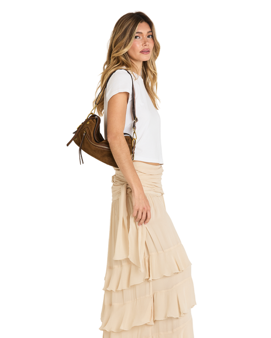 A woman in a white t-shirt and a beige ruffled tiered maxi skirt is shown from the side, carrying a brown leather shoulder bag against a solid white background.
