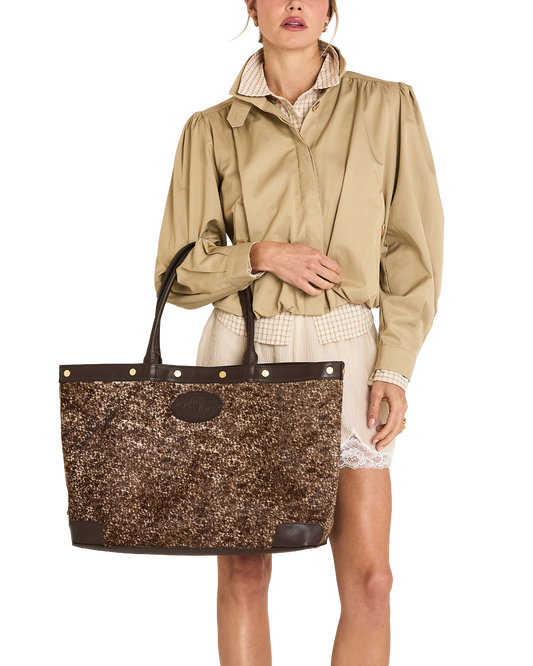 A model wearing a beige trench coat and cream shorts holds a large brown glittery tote bag with dark brown leather trim and handles against a plain white background.