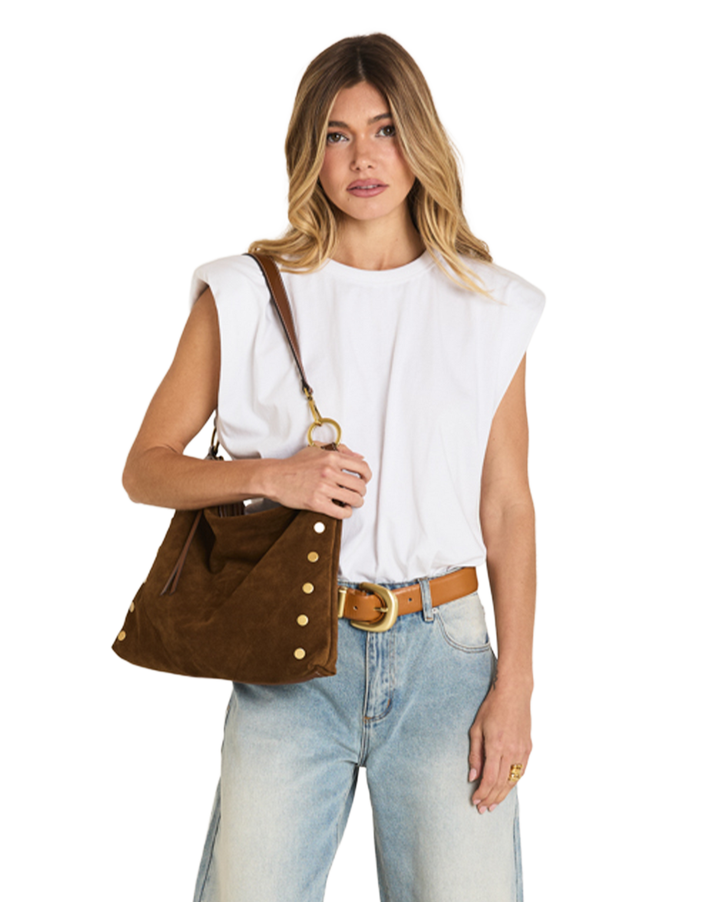 A woman modeling a brown suede shoulder bag with gold-tone studs and a leather strap, styled with a white sleeveless top, light blue jeans, and a tan belt against a plain white background.