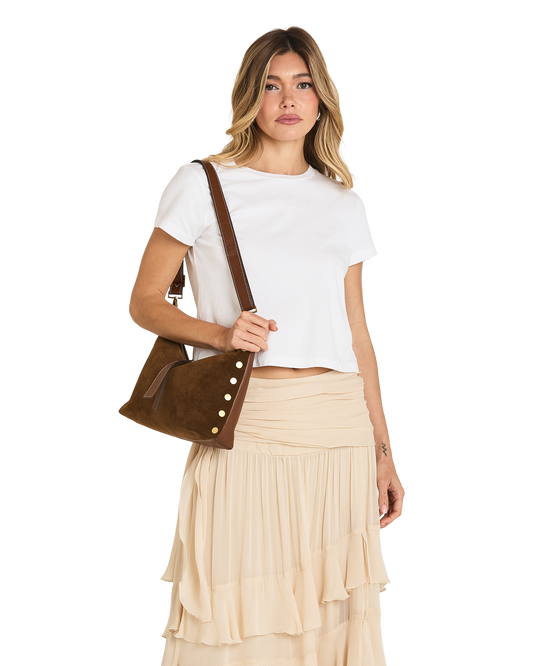 A woman modeling a brown suede crossbody bag with decorative gold-tone buttons, styled with a white crop top and a beige pleated skirt against a plain white background.
