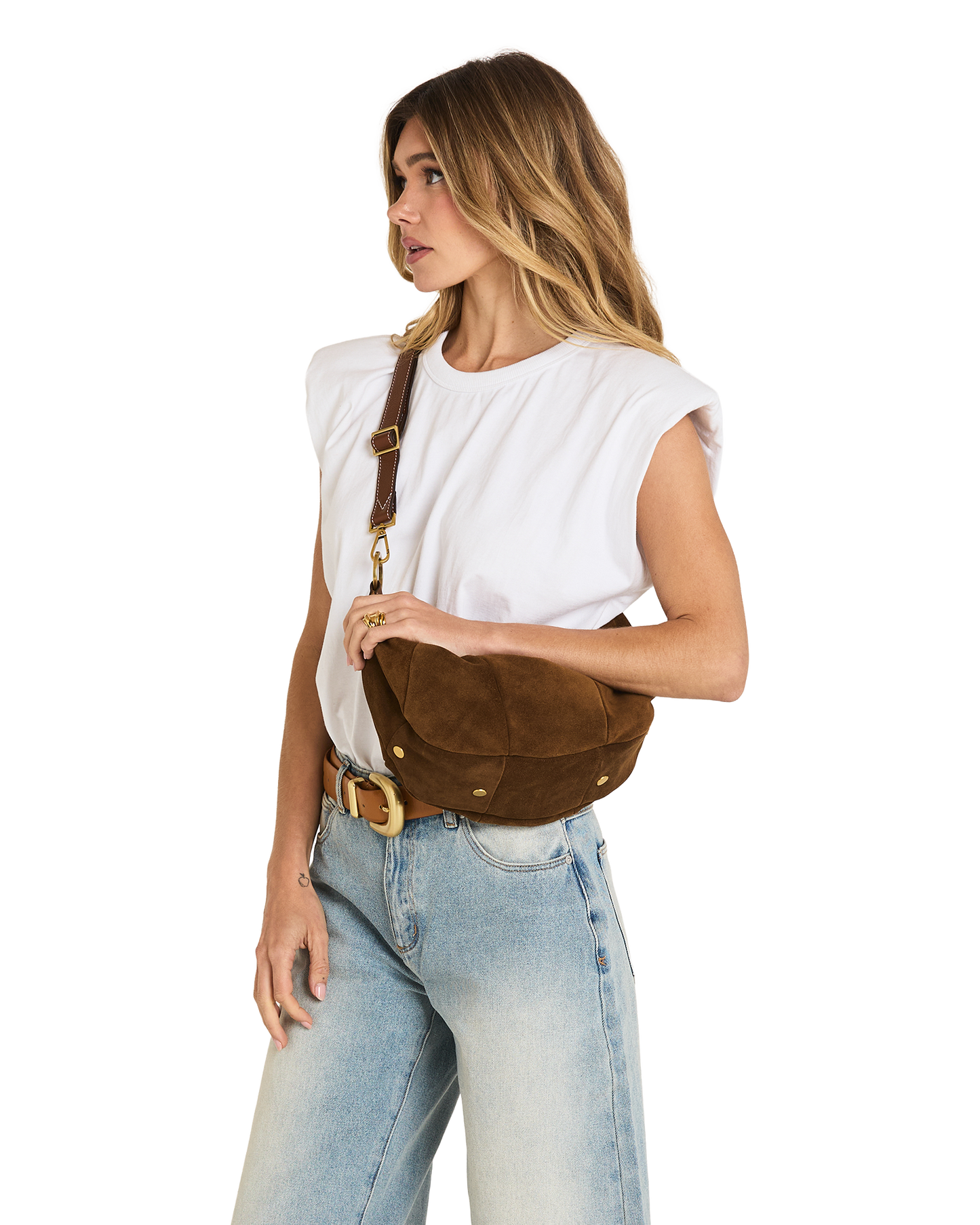 A woman wearing a white sleeveless top and light blue jeans models a brown suede crescent-shaped crossbody bag with gold-tone hardware against a plain white background.
