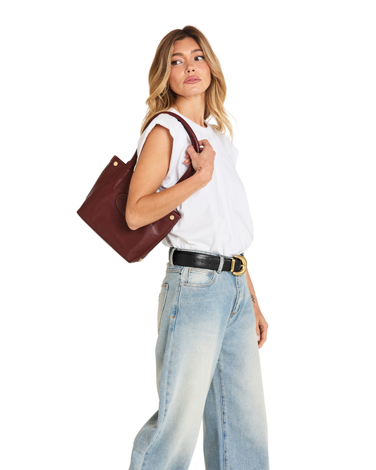 A woman wearing a white sleeveless top, light blue wide-leg jeans, and a black belt with a gold buckle, carrying a dark brown leather tote bag over her shoulder against a plain white background.