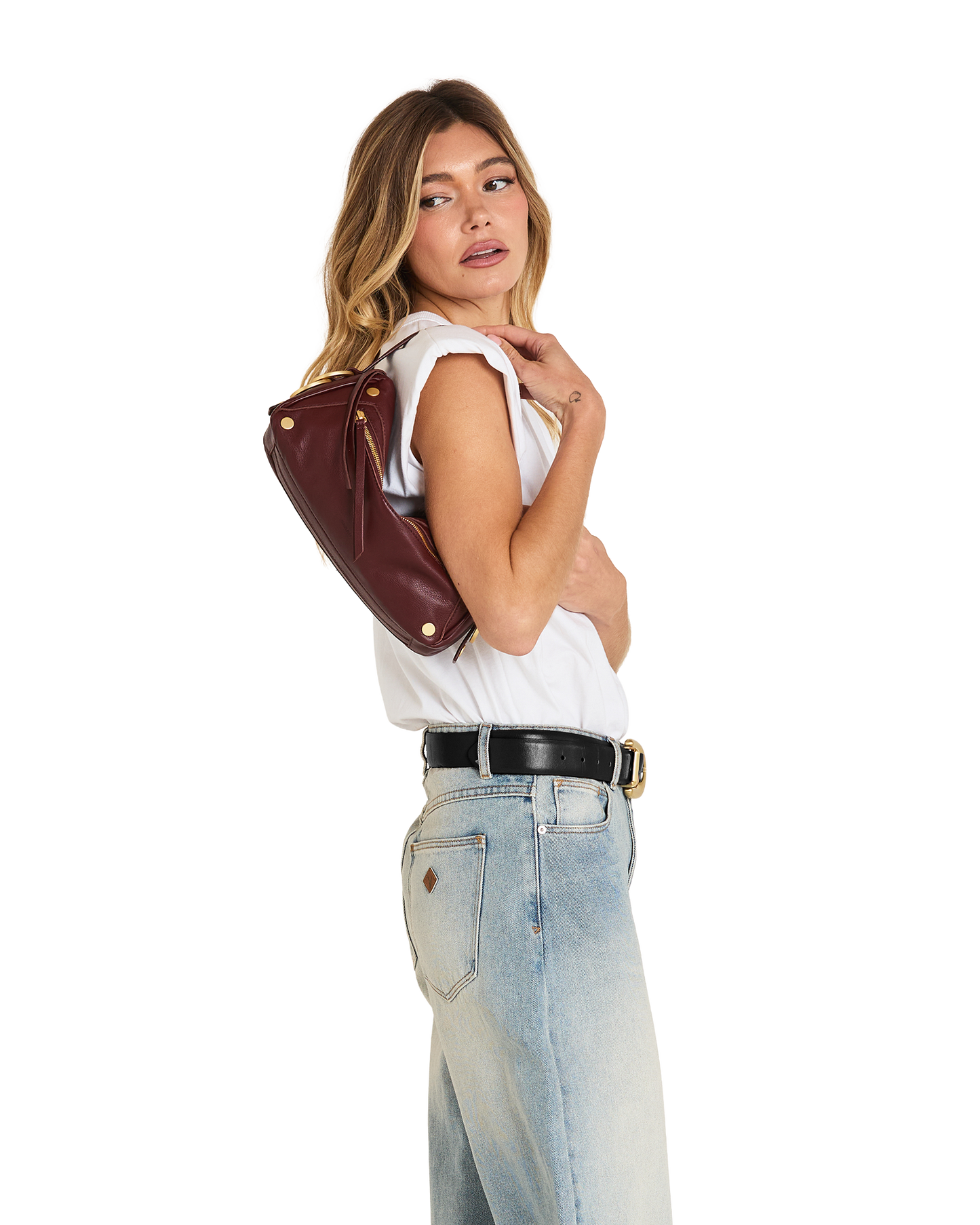 A woman posing in a white sleeveless top, light wash jeans, and a black belt, carrying a dark brown leather shoulder bag with gold-tone hardware against a plain white background.
