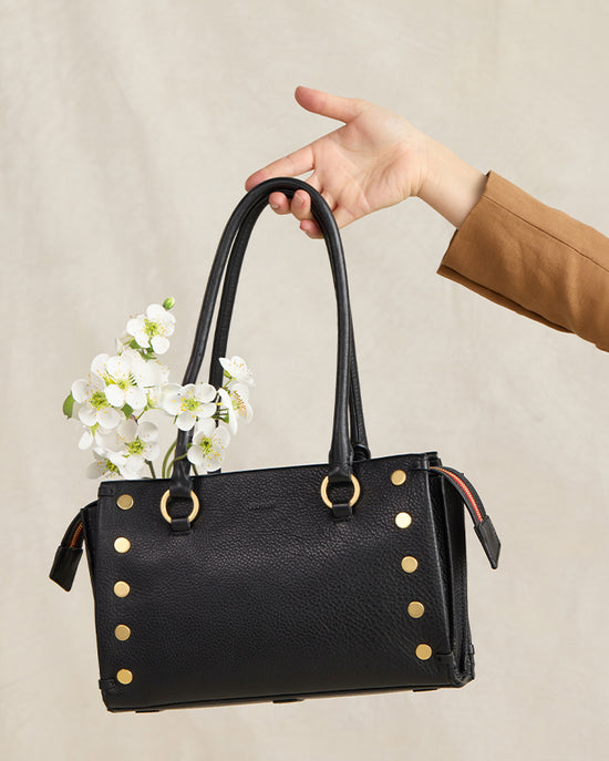 Image of hand holding Allen Lrg in brushed gold red zip in front of light tan backdrop with flowers coming out of the bag.