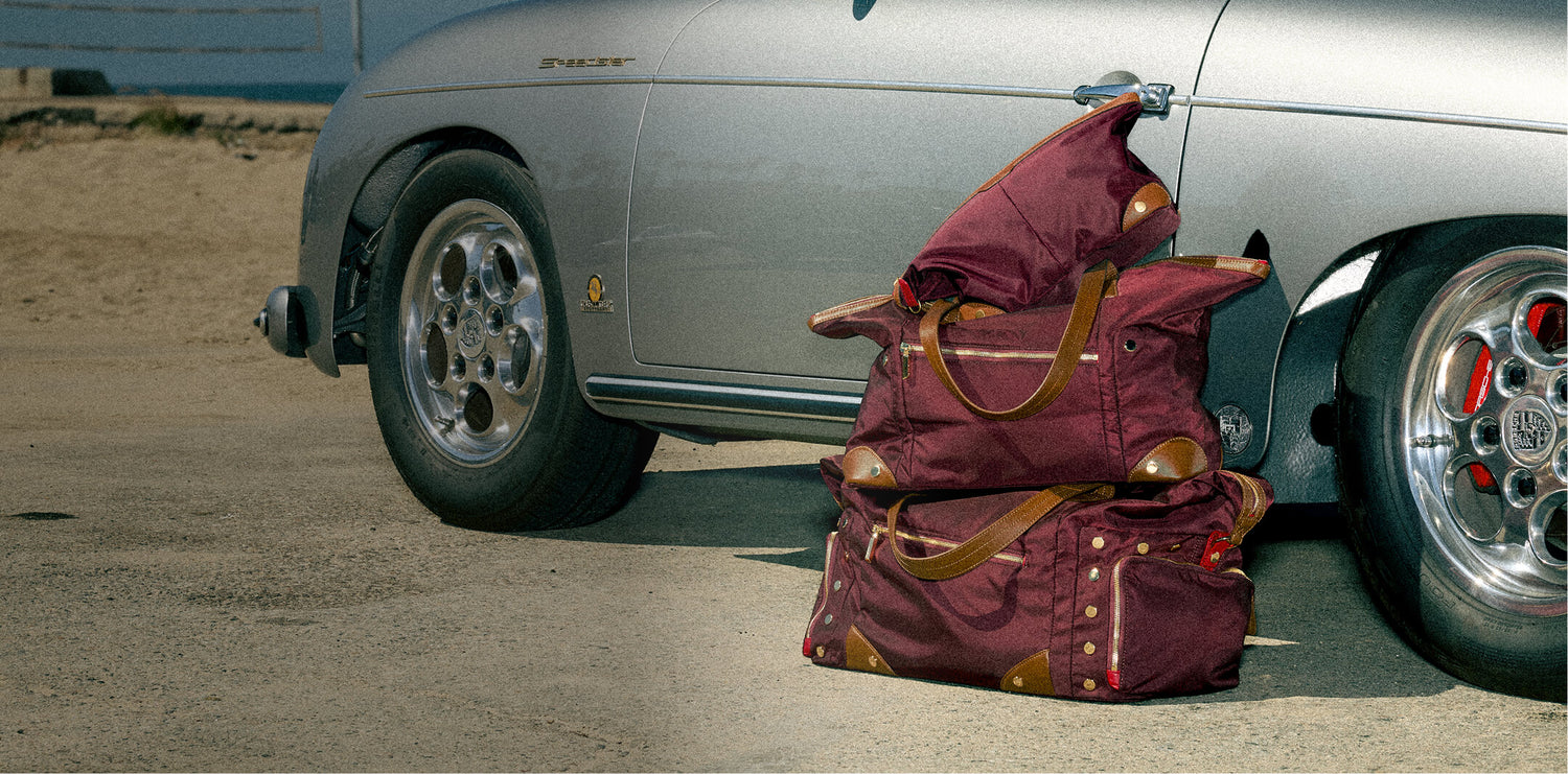 A stack of three burgundy duffel bags with tan leather handles and gold-toned hardware sits beside a silver classic car on a paved outdoor surface.