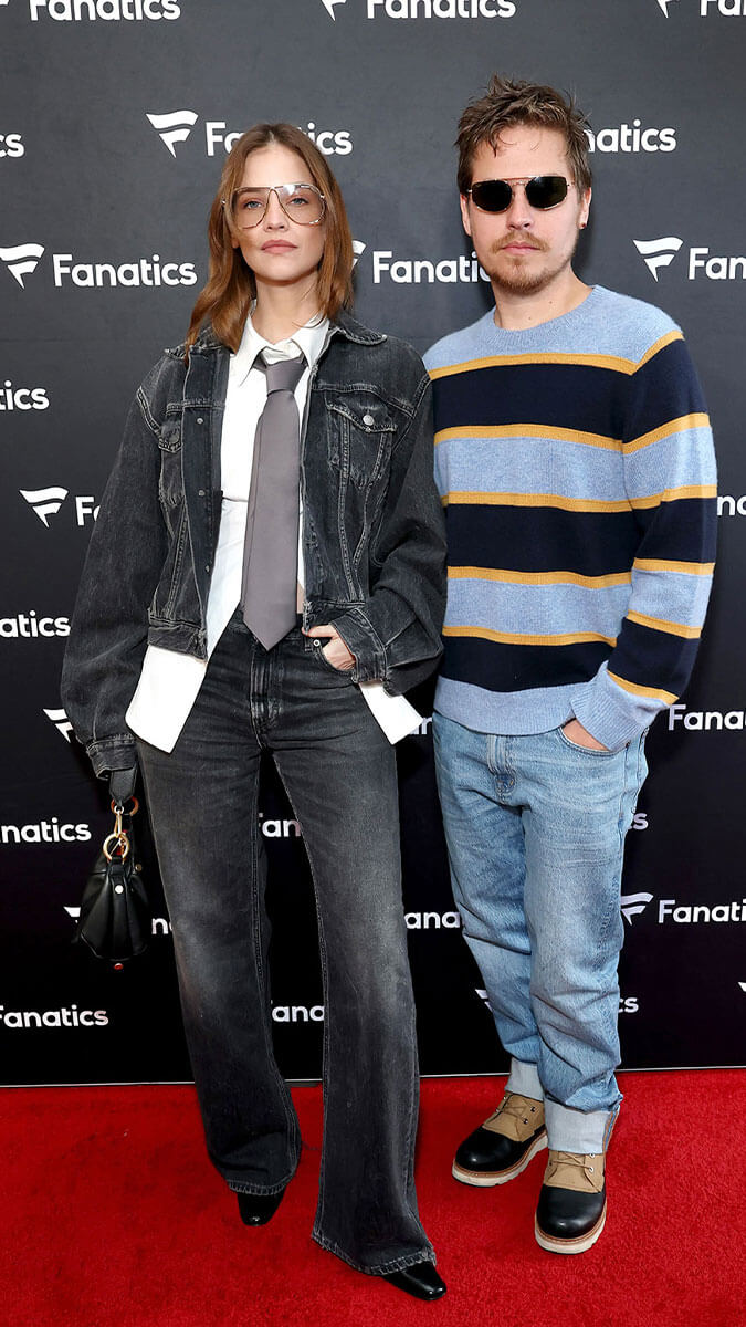 A couple posing on a red carpet in front of a black Fanatics backdrop. The woman wears a black denim jacket and flare jeans over a white shirt and gray tie, while the man wears a blue, navy, and yellow striped sweater, light blue jeans, and brown shoes.